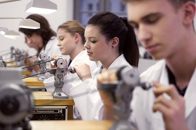 Datei:05 watchmaking apprentices at turning machines.jpg
